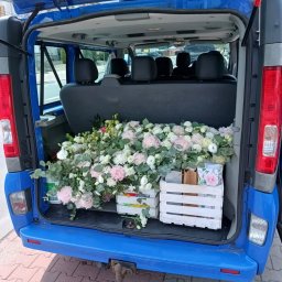 BIKE-BUS KAMIL MATUSIAK - Niebieski bus z otwartym bagażnikiem wypełnionym kwiatami w białych skrzynkach. Widoczne tylne siedzenia i fragment ulicy w tle. Dostawa kwiatów na uroczystość.