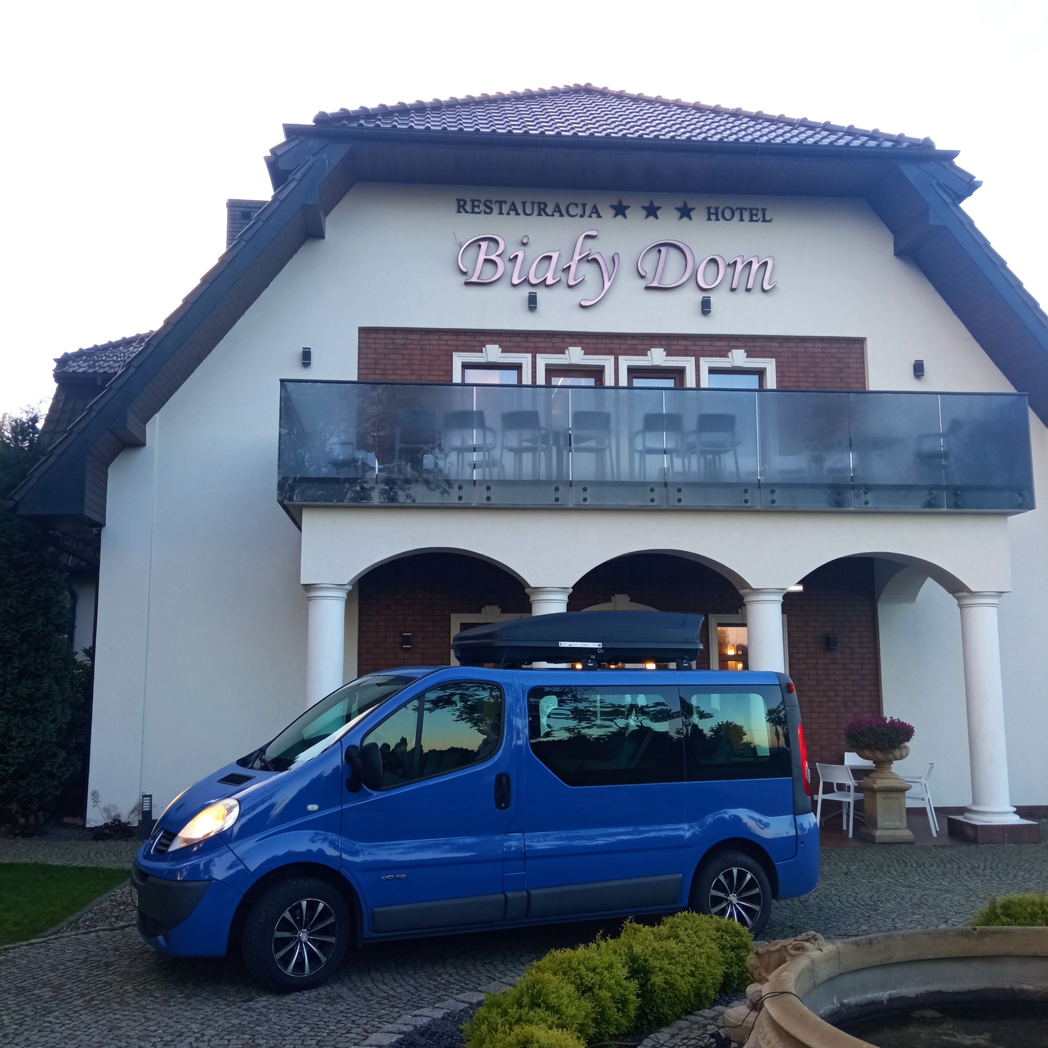 Niebieski bus z bagażnikiem dachowym zaparkowany przed budynkiem z napisem 'Restauracja Hotel Biały Dom'.