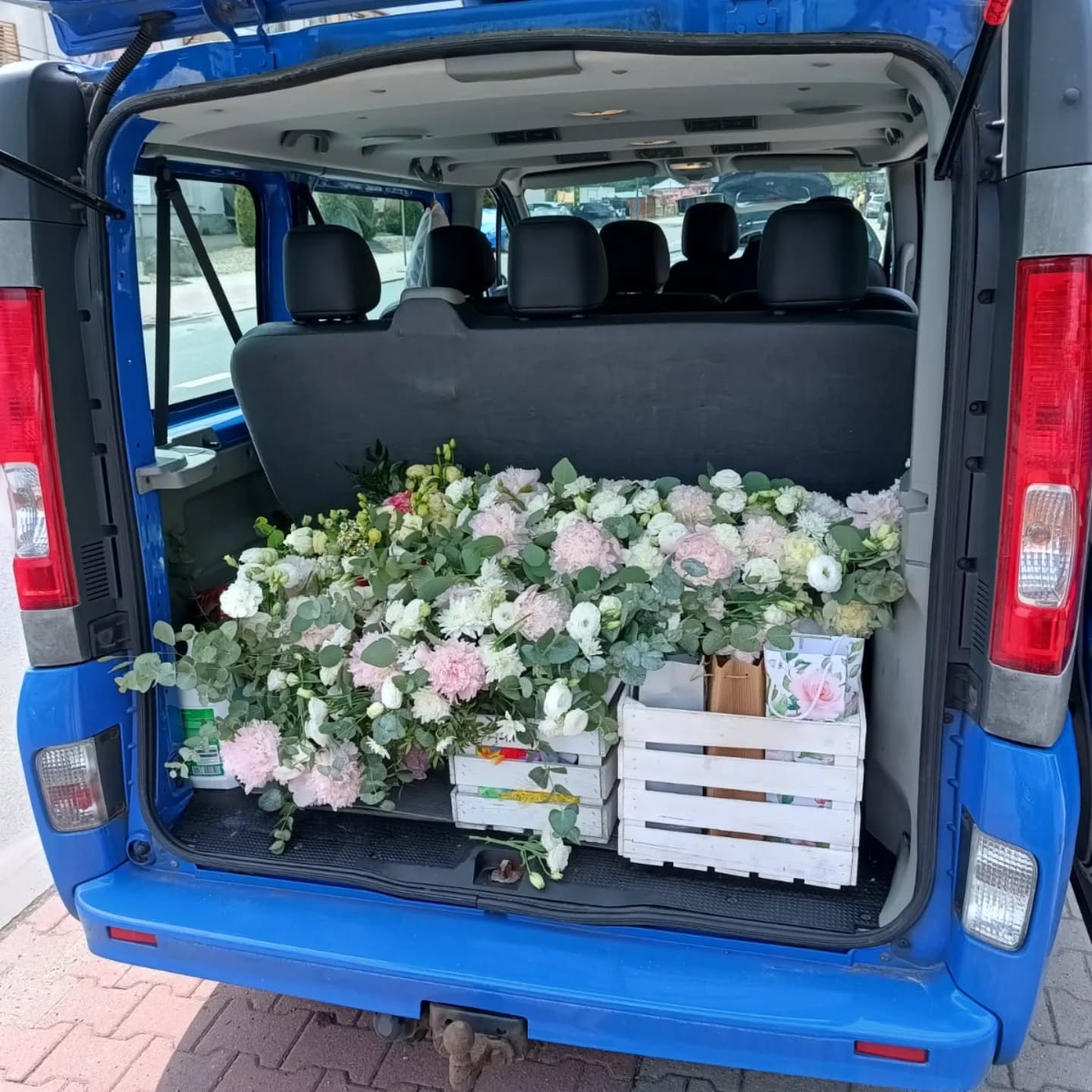 Niebieski bus z otwartym bagażnikiem wypełnionym kwiatami w białych skrzynkach. Widoczne tylne siedzenia i fragment ulicy w tle. Dostawa kwiatów na uroczystość.