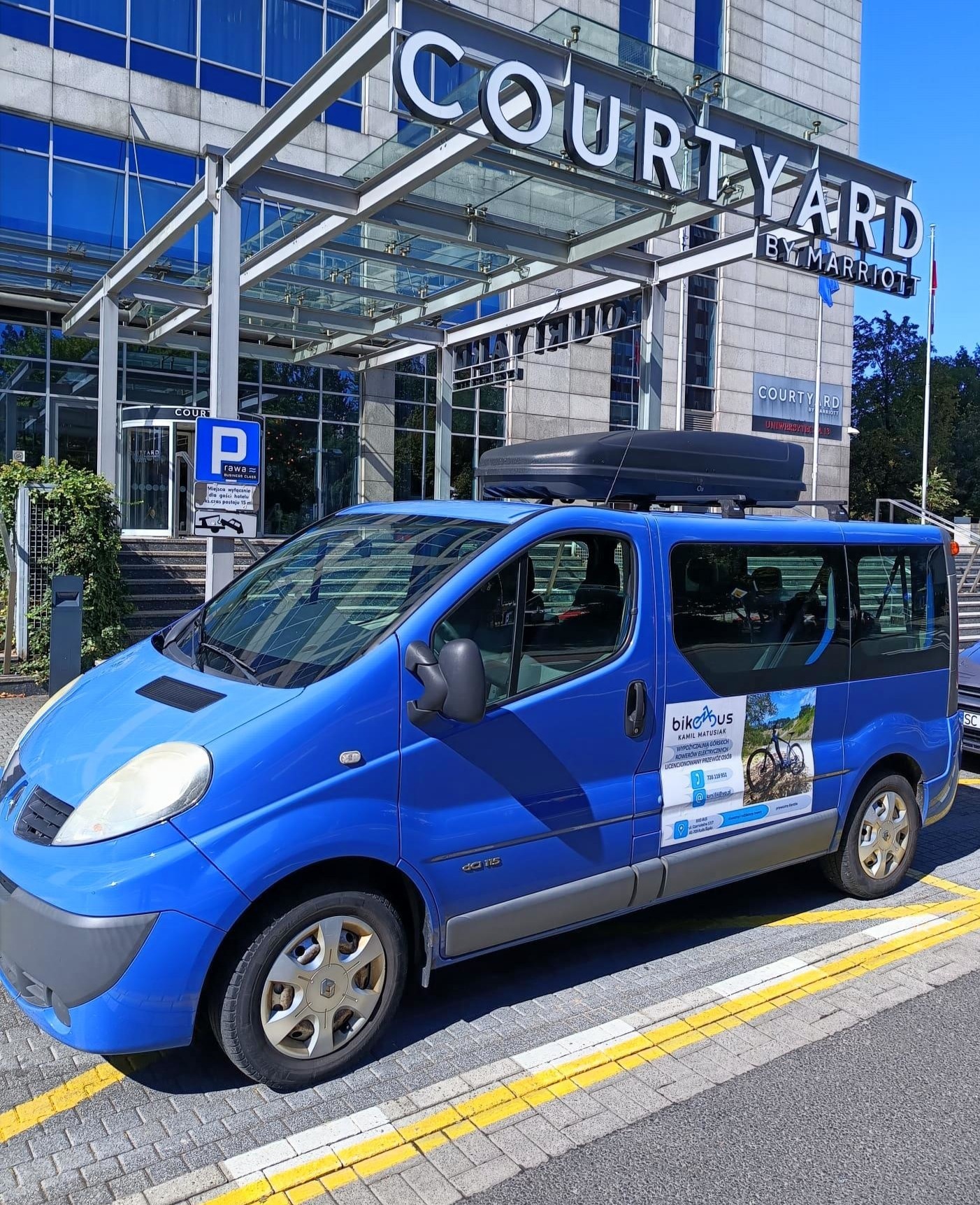 Niebieski bus z bagażnikiem dachowym i reklamą 'bikebus' na parkingu przed hotelem Courtyard by Marriott w słoneczny dzień. Widoczny znak parkingowy.