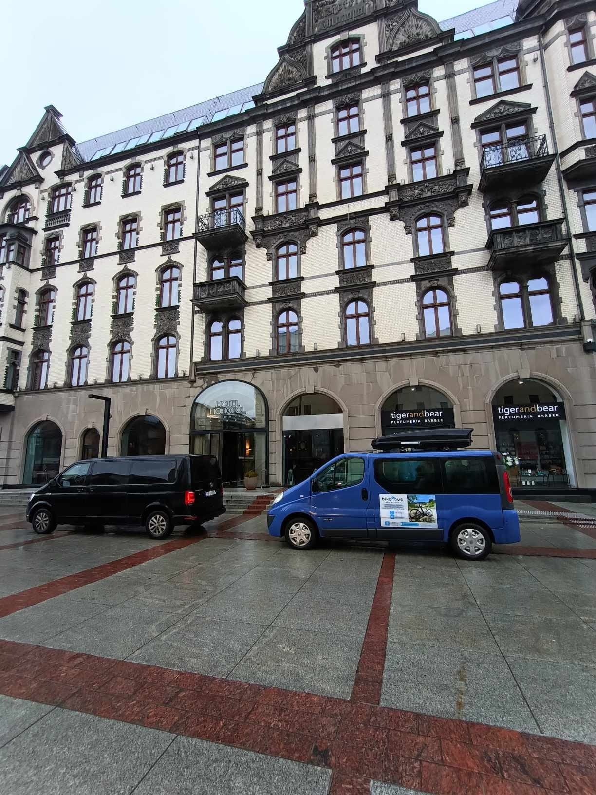Dwa busy (czarny i niebieski z bagażnikiem dachowym) zaparkowane przed hotelem o architekturze secesyjnej. Na niebieskim busie widoczne naklejki reklamowe.