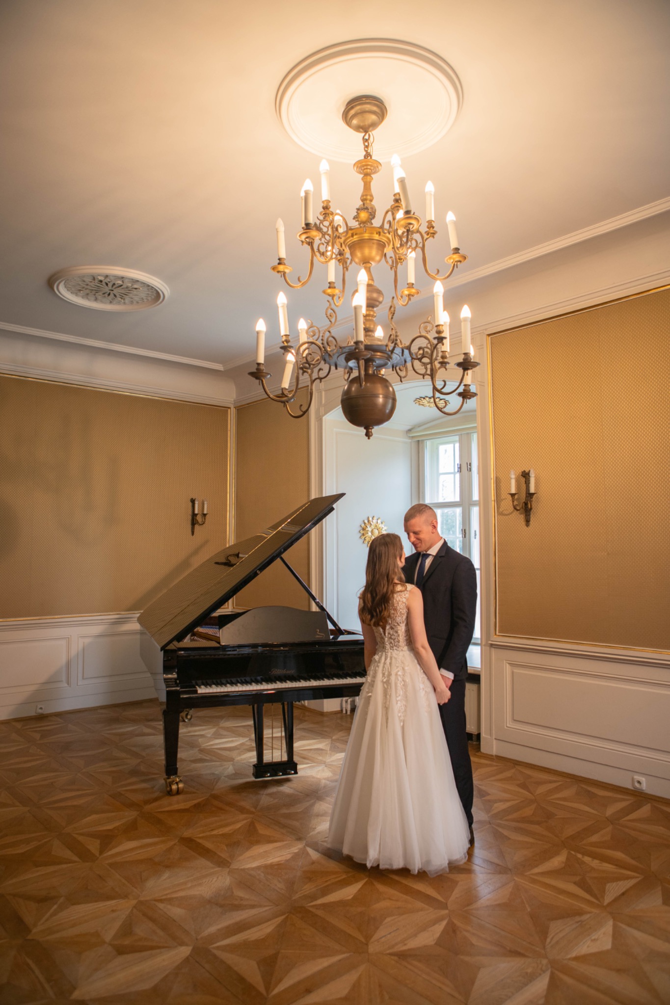 Para młoda trzymająca się za ręce w eleganckim wnętrzu z fortepianem i żyrandolem. Ujęcie z tyłu, suknia ślubna z koronkowymi detalami, ciemny garnitur.