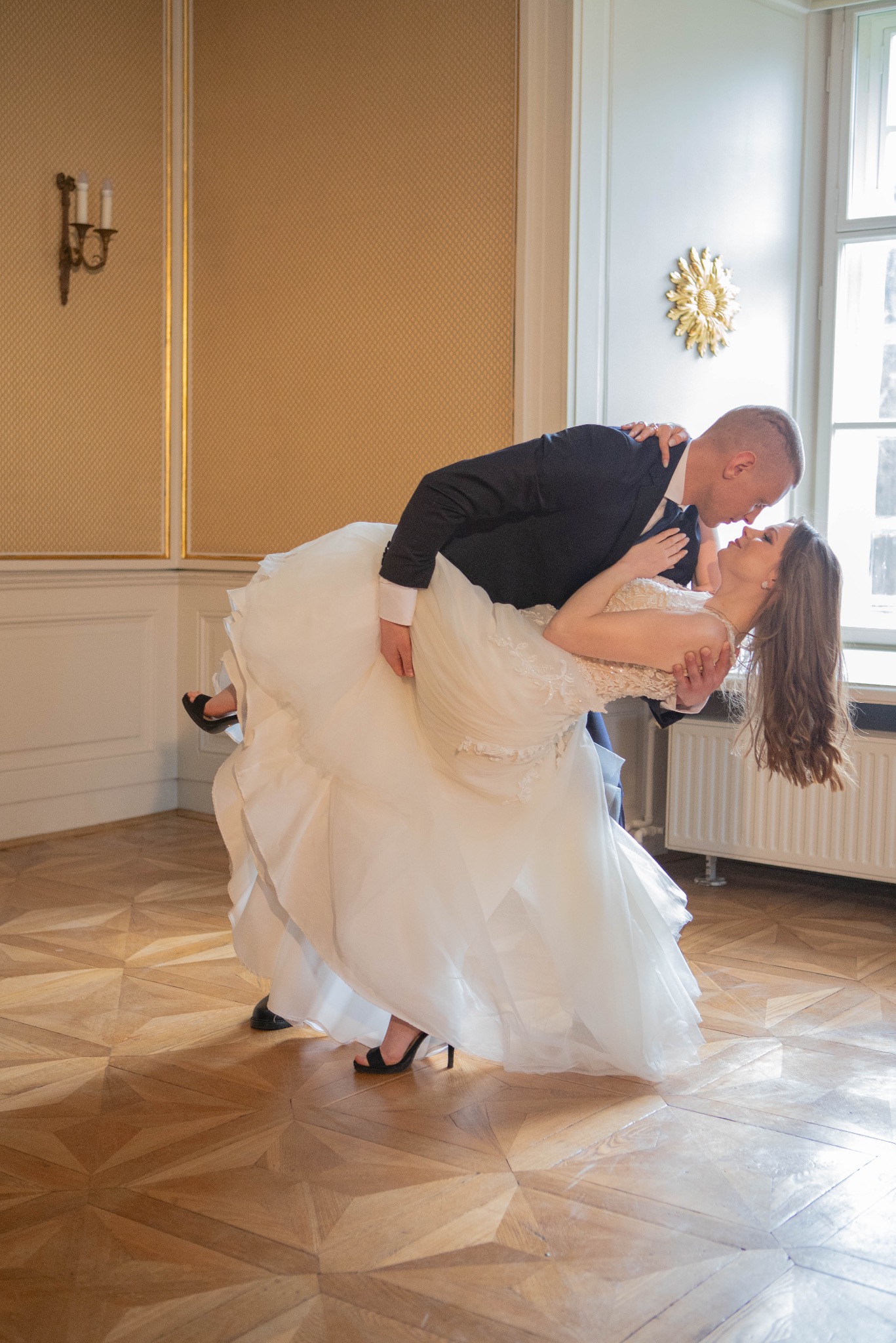 Para młoda w tańcu w eleganckim wnętrzu. Panna młoda w sukni z koronkową górą, pan młody w ciemnym garniturze. Parkiet z wzorem, dekoracje ścienne.