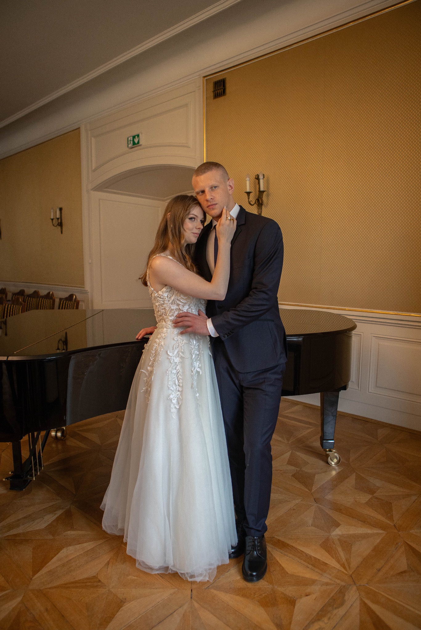 Elegancka sesja ślubna: para młoda w objęciach przy czarnym fortepianie w klasycznym wnętrzu z parkietem i złoconymi ścianami. Ona w sukni z koronką, on w garniturze.