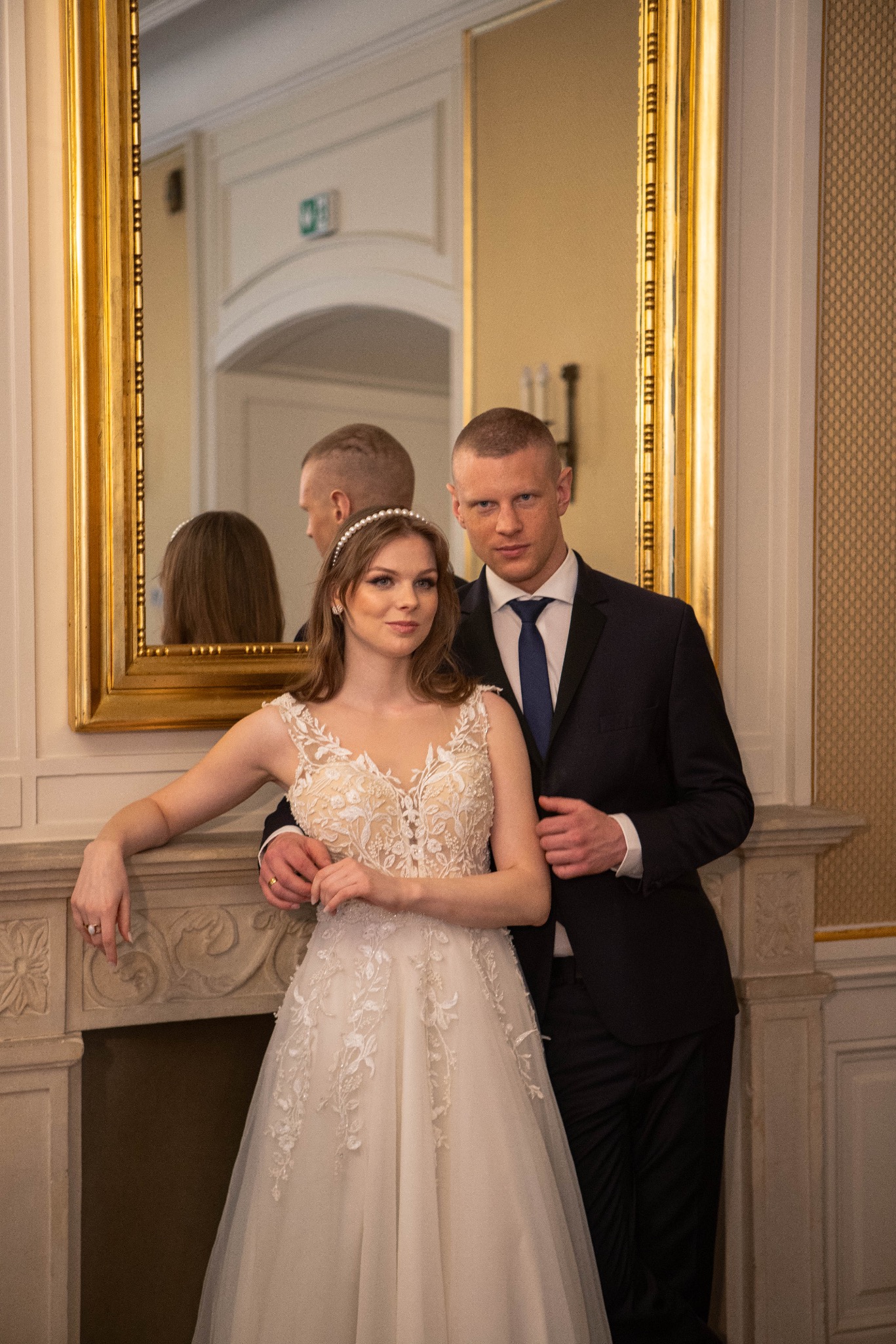 Elegancka para młoda w strojach ślubnych pozuje przy kominku i lustrze w złotej ramie. Ona w koronkowej sukni, on w granatowym garniturze z niebieskim krawatem.