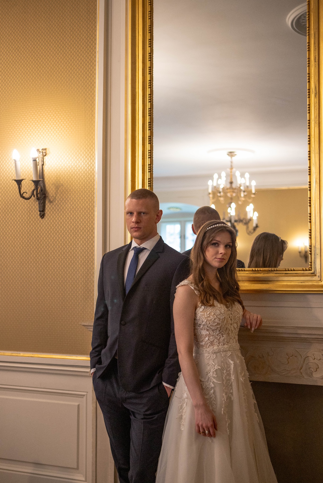 Elegancka para młoda w formalnych strojach pozuje w luksusowym wnętrzu z lustrem w złotej ramie, odbijającym żyrandol i fragment pomieszczenia.