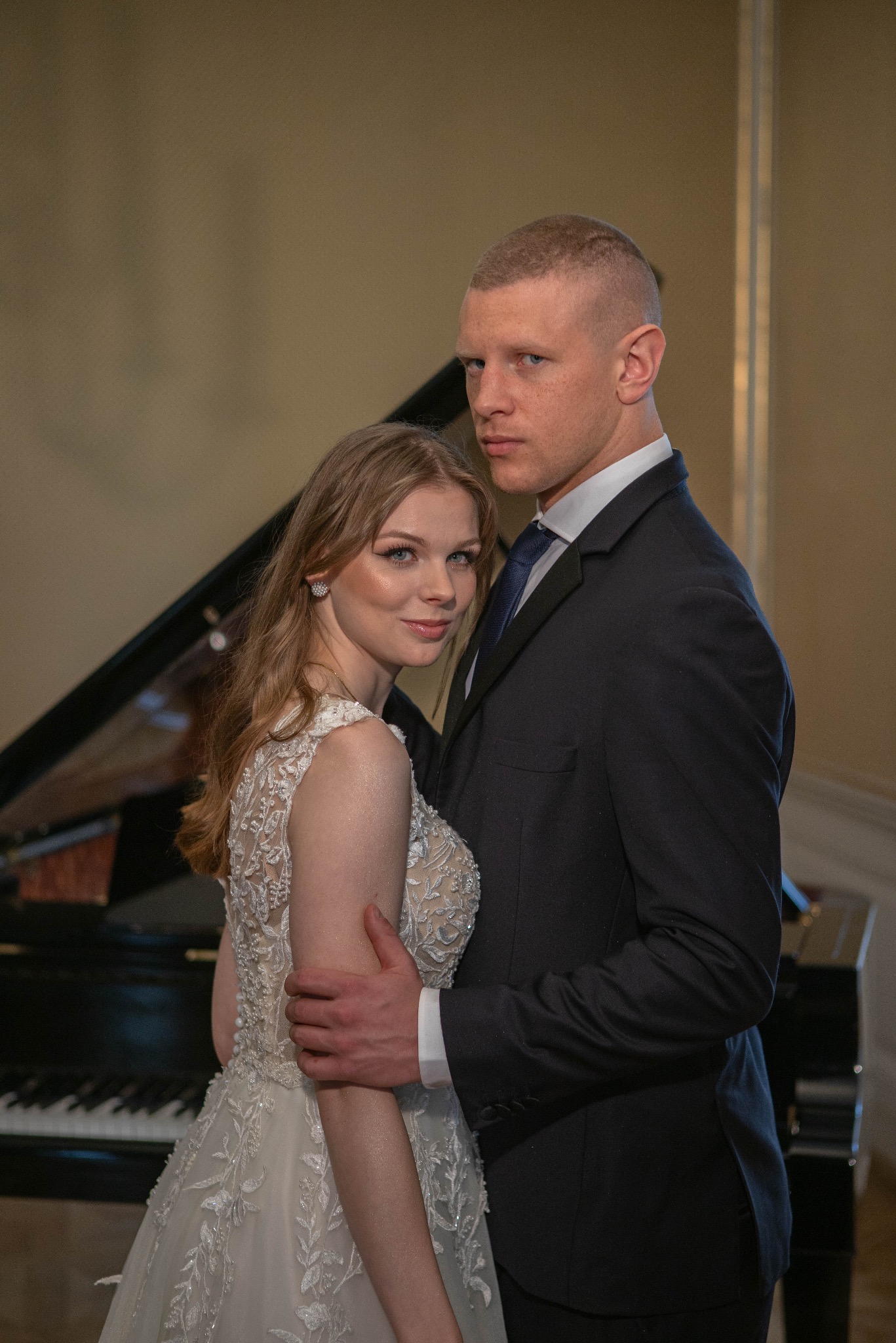 Elegancka para młoda w objęciach na tle czarnego fortepianu. Ona w sukni z koronkowymi zdobieniami, on w granatowym garniturze i krawacie. Subtelne oświetlenie podkreśla uroczysty charakter.
