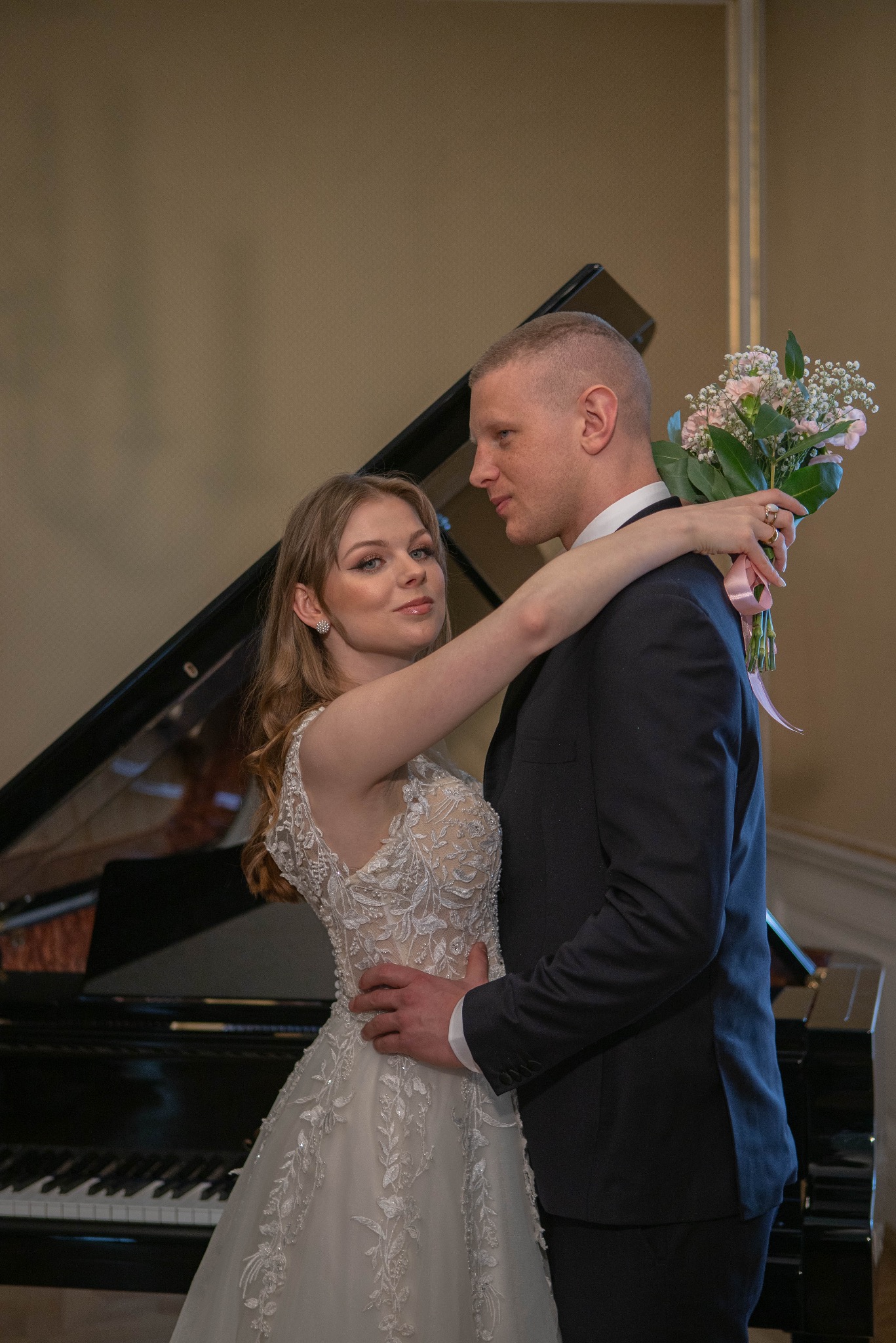 Para młoda w objęciach na tle czarnego fortepianu. Panna młoda w sukni z koronkowymi aplikacjami trzyma bukiet różowych kwiatów.