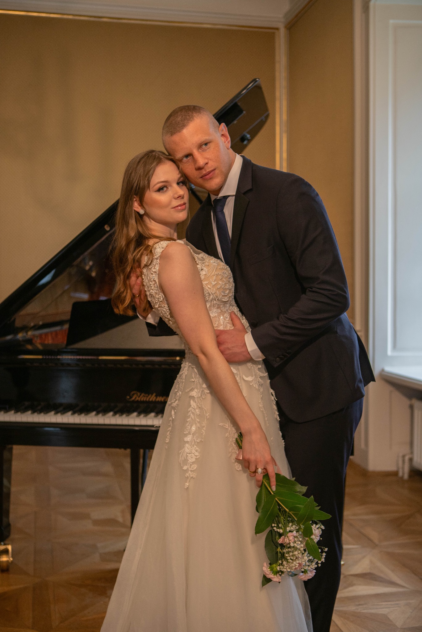 Elegancka para młoda w objęciach na tle czarnego fortepianu Blüthner. Panna młoda w sukni z koronkowymi aplikacjami trzyma bukiet. Pan młody w granatowym garniturze i krawacie.