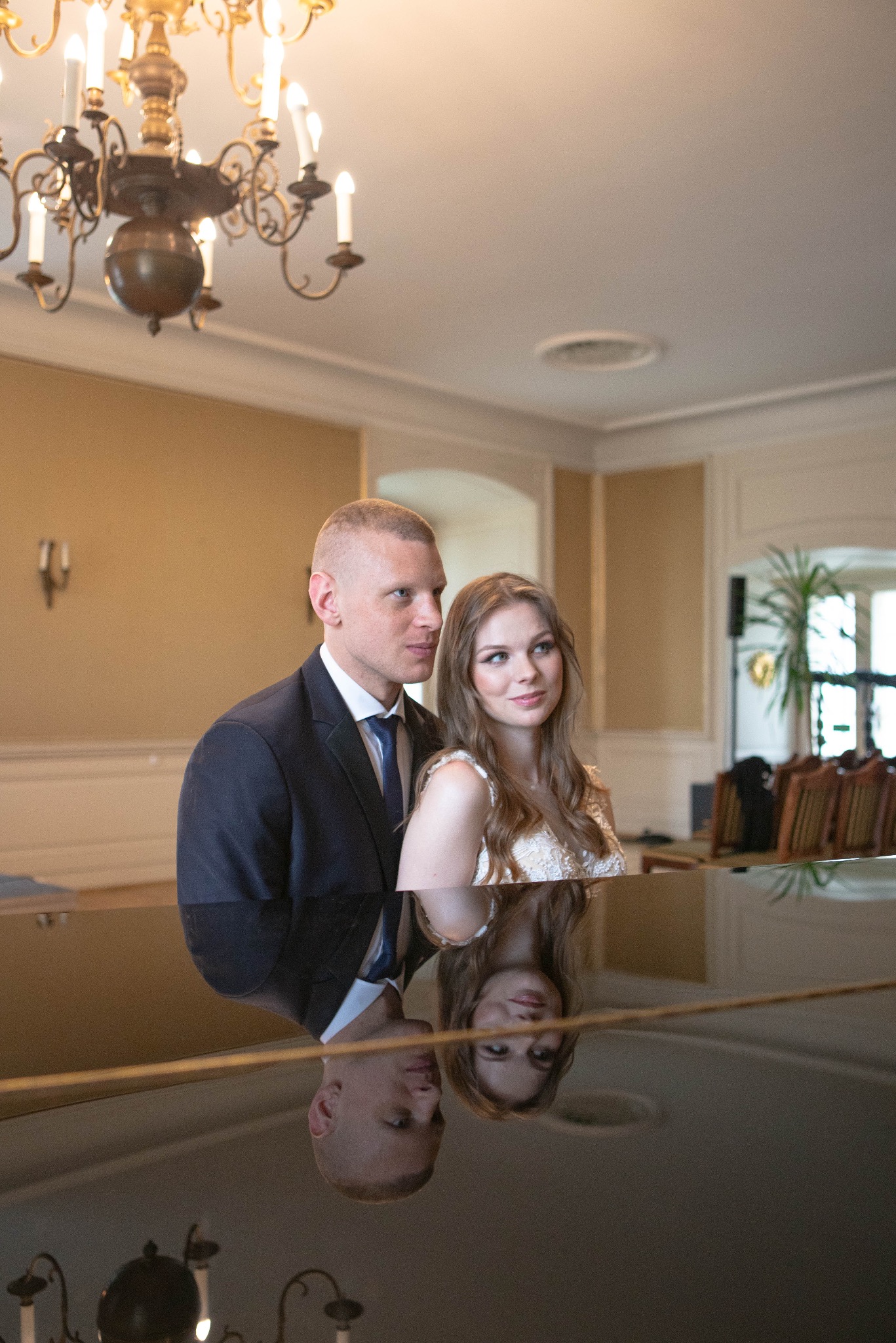 Elegancka para młoda pozuje w przestronnym wnętrzu z żyrandolem, odbijając się w lakierowanej powierzchni fortepianu. Ujęcie z dołu podkreśla uroczysty charakter.