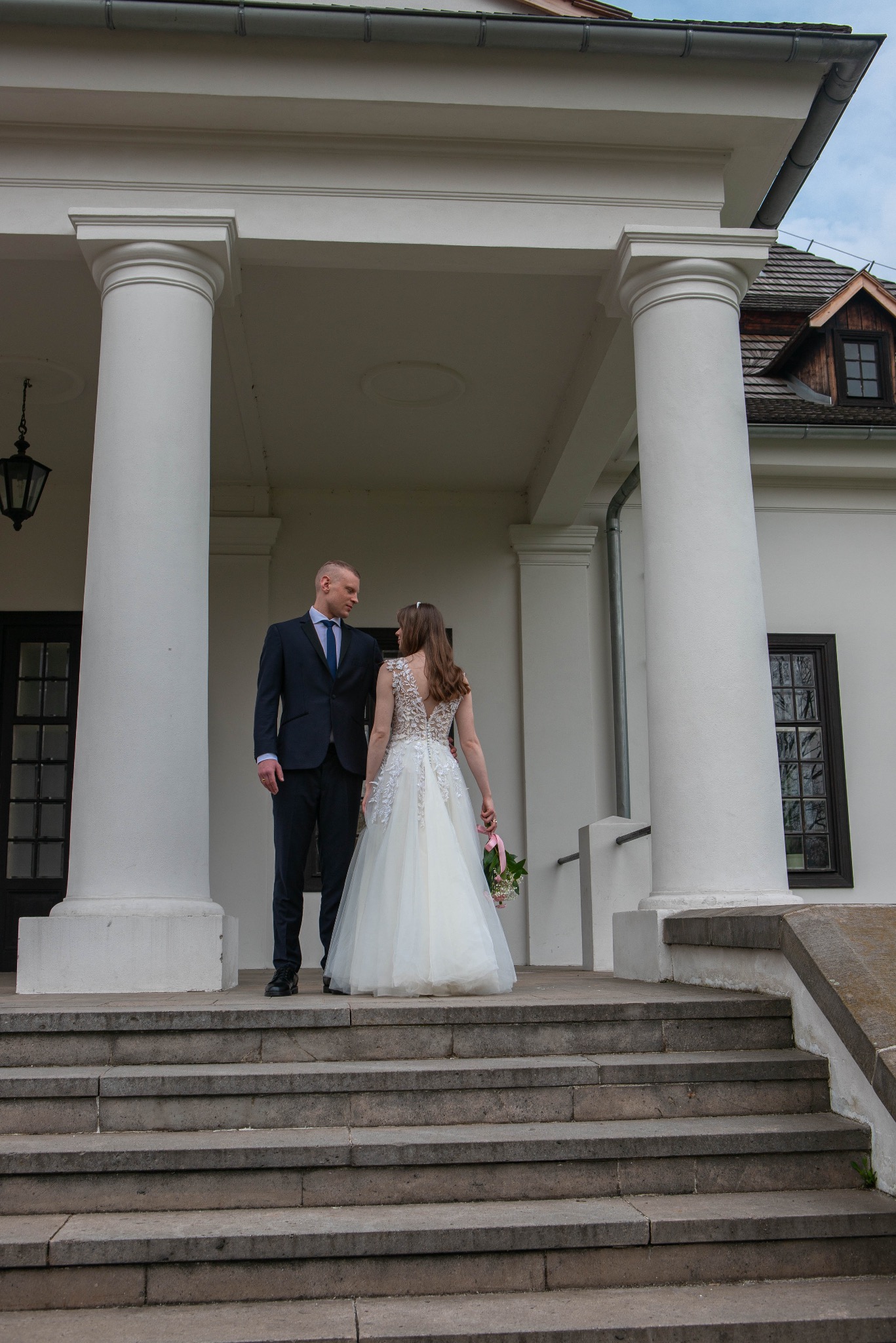 Para młoda na schodach klasycystycznego budynku z kolumnami. Panna młoda w sukni z odkrytymi plecami, pan młody w granatowym garniturze. Romantyczny kadr.