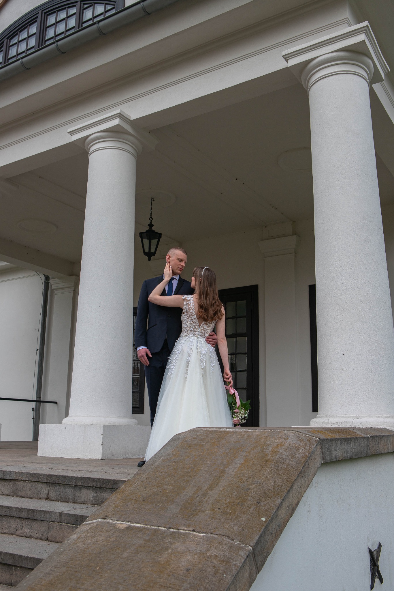 Para młoda w eleganckich strojach pozuje na tle kolumnady z latarnią. Panna młoda w sukni z koronkowymi detalami, pan młody w ciemnym garniturze. Romantyczna sceneria.
