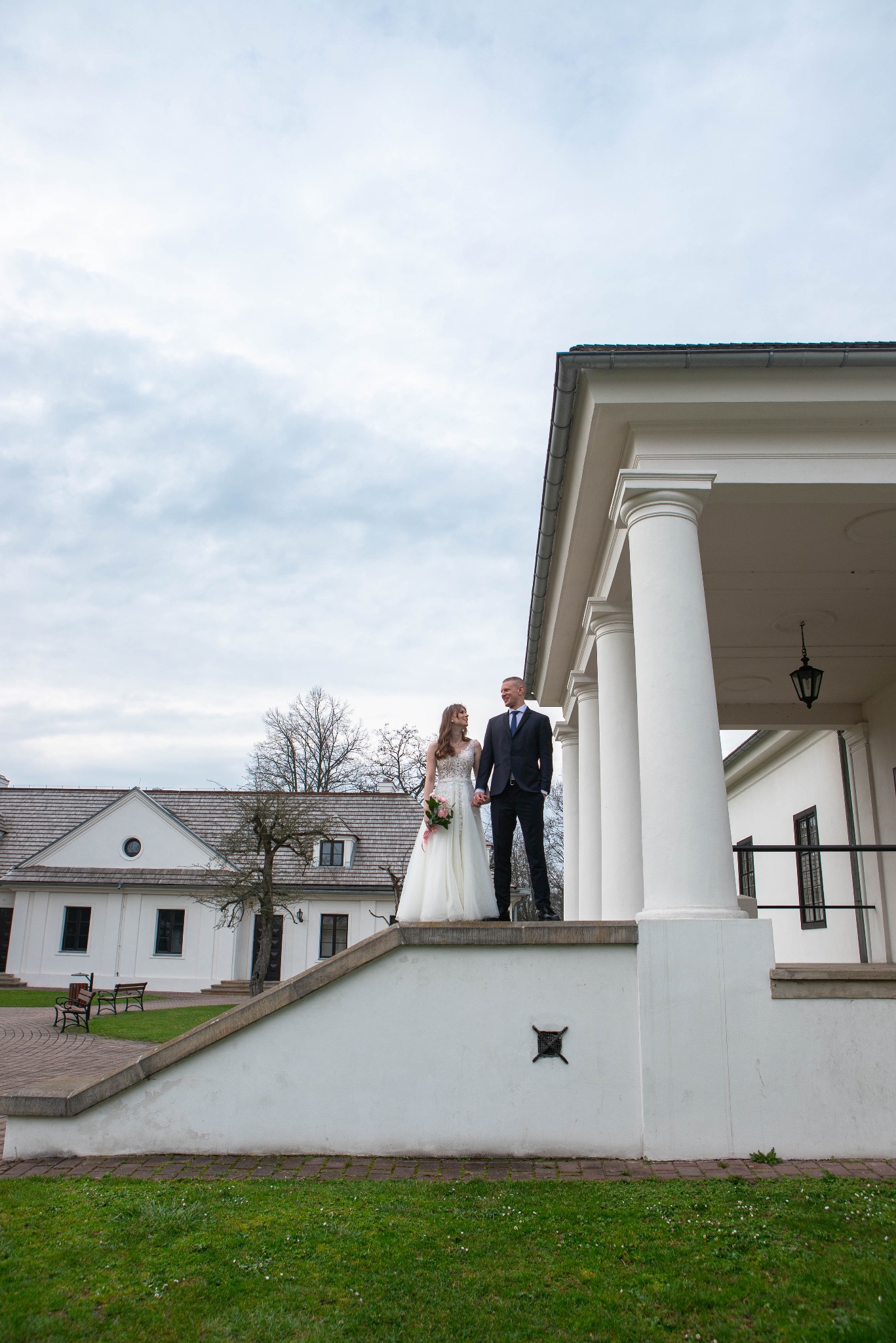 Para młoda w Trzemeśni pozuje na tle klasycystycznego budynku z kolumnami. Panna młoda trzyma bukiet, a pan młody ubrany jest w garnitur. Pochmurne niebo.