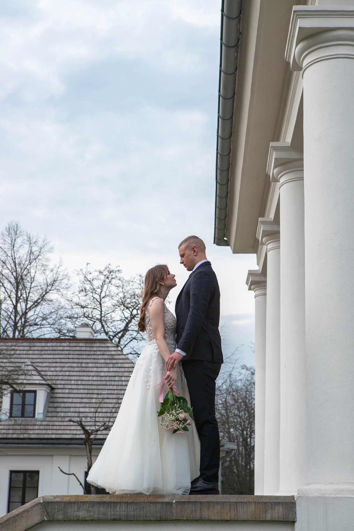 Para młoda w eleganckich strojach stoi na tle klasycystycznej architektury z kolumnami i budynkiem w tle. Panna młoda trzyma bukiet z różową wstążką.