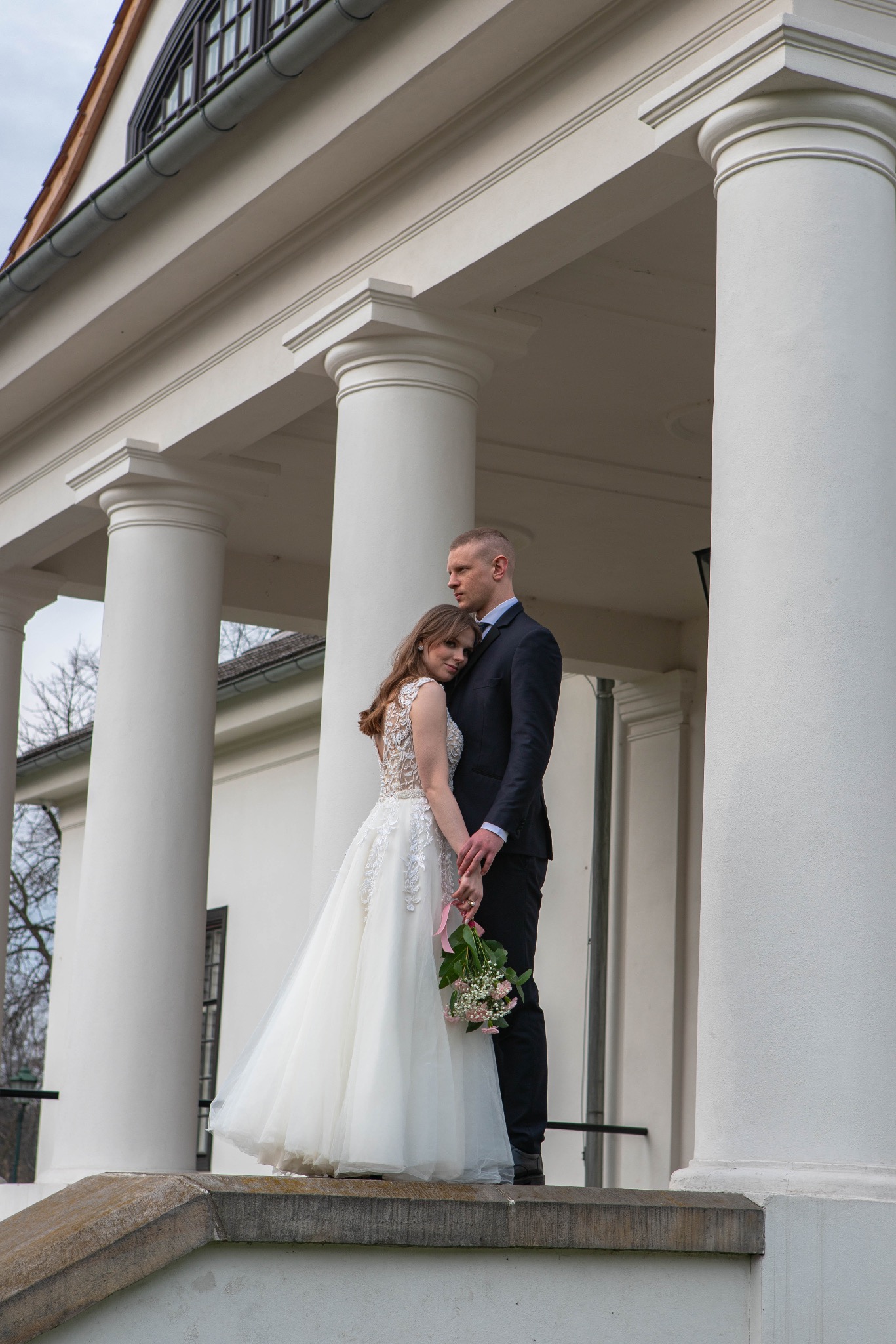 Para młoda w eleganckich strojach pozuje na tle klasycystycznej architektury z kolumnami. Panna młoda trzyma bukiet, a pan młody obejmuje ją czule.