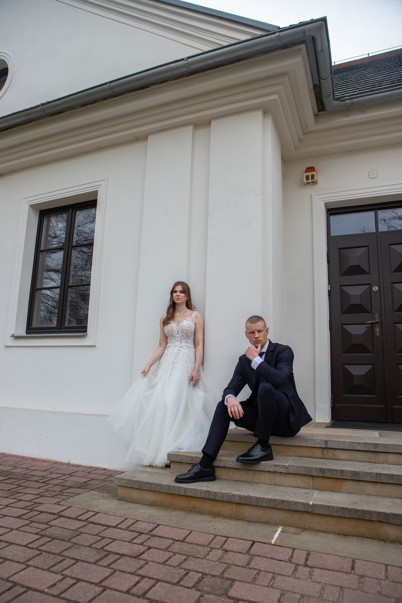 Elegancka sesja ślubna: Panna młoda w koronkowej sukni opiera się o białą ścianę, pan młody w granatowym garniturze siedzi na schodach, budynek w tle.