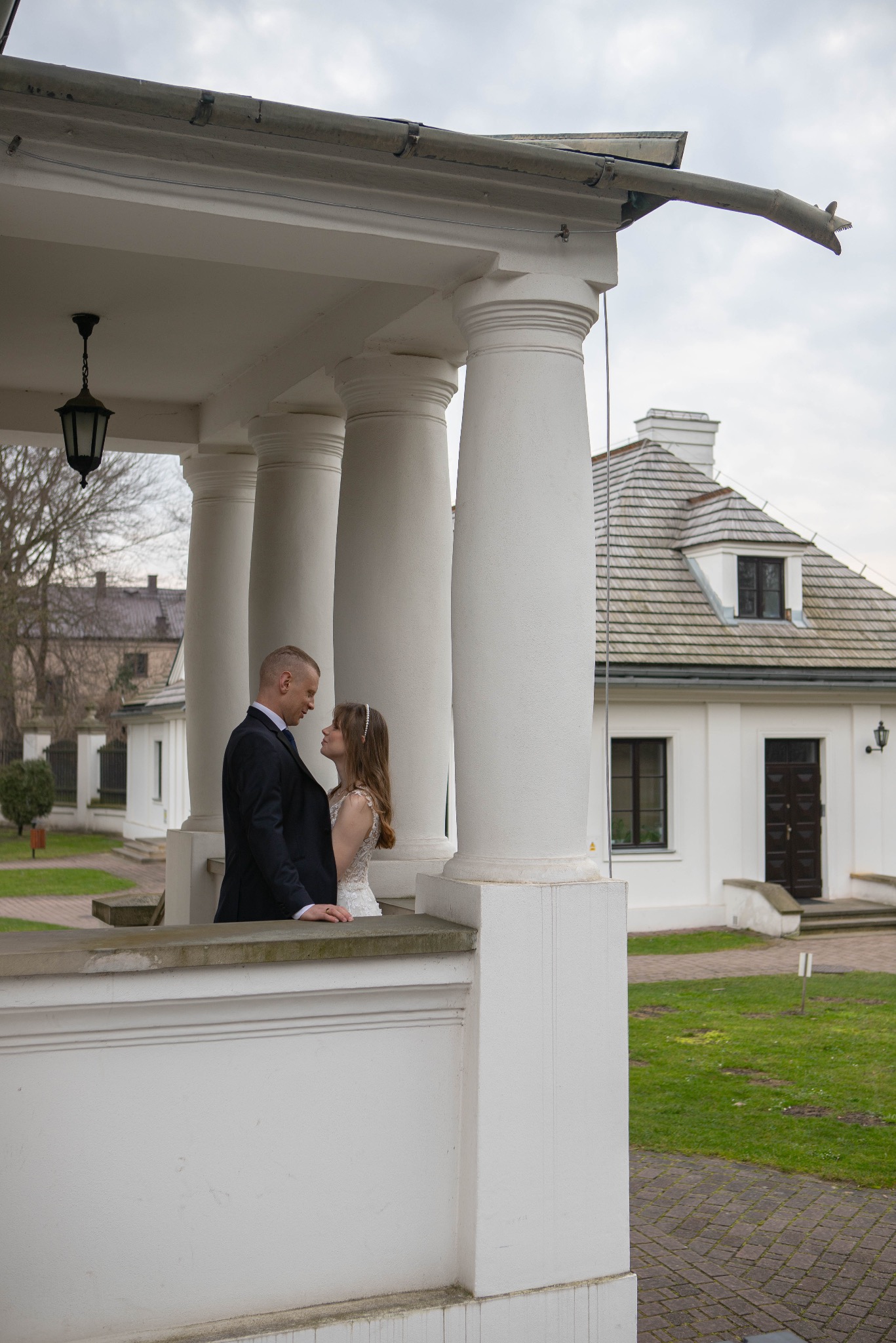 Para młoda w objęciach na werandzie z kolumnami, w tle biały budynek z szarym dachem. Ujęcie z dołu, podkreślające architekturę i intymność chwili.