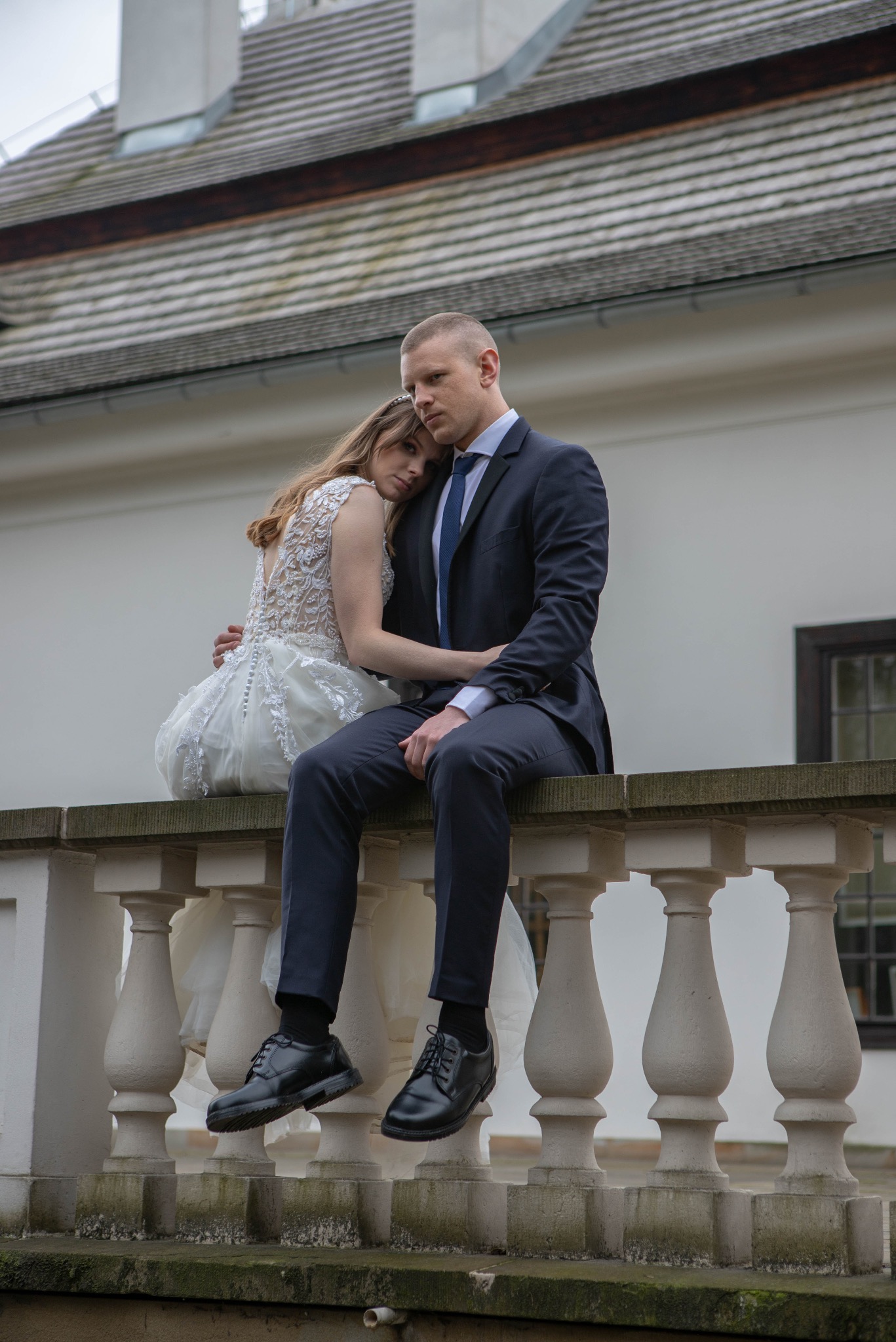 Para młoda w eleganckich strojach siedzi na kamiennej balustradzie na tle białego budynku z szarym dachem. Panna młoda opiera głowę na ramieniu pana młodego.