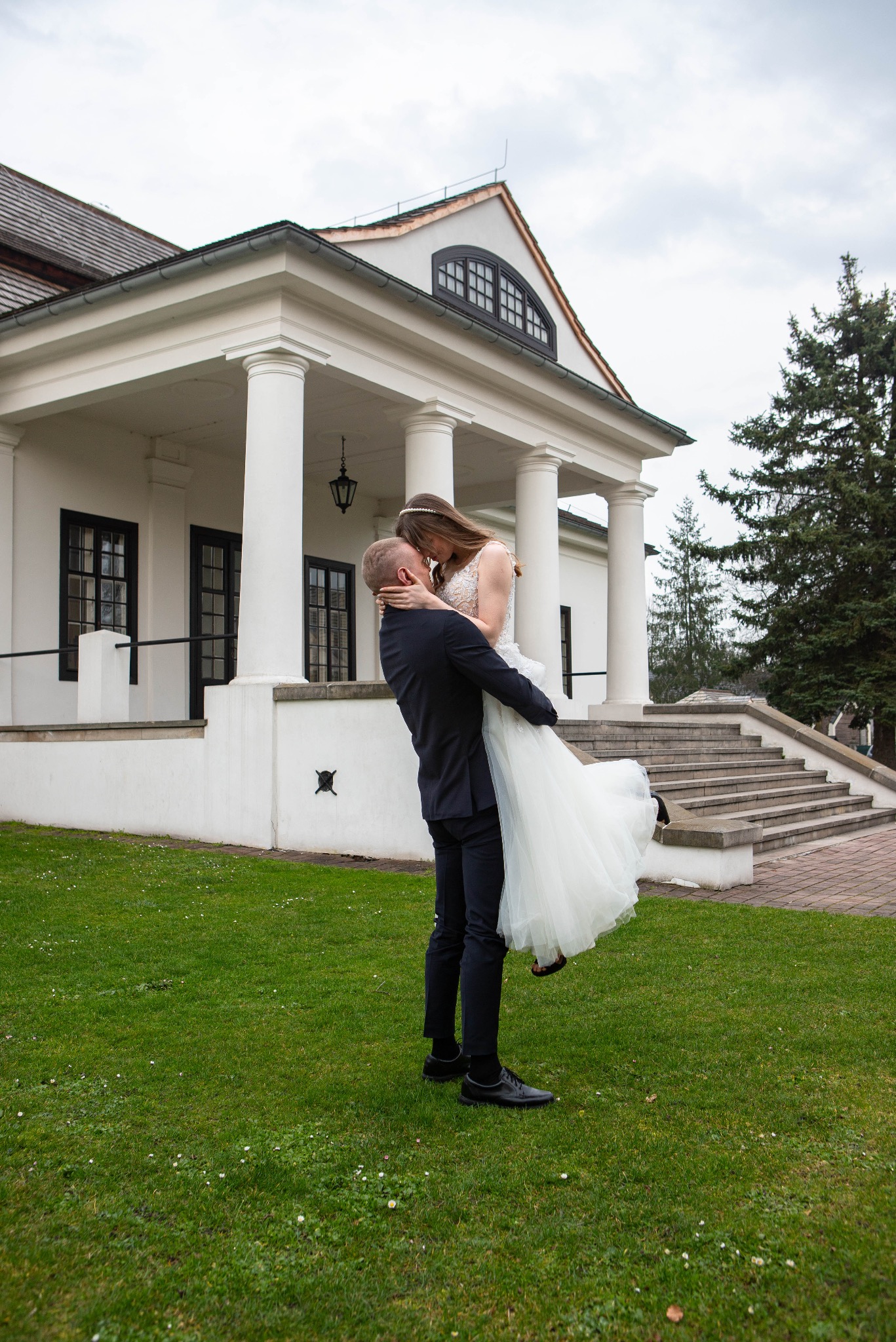 Uśmiechnięty pan młody trzyma pannę młodą w białej sukni na tle klasycystycznego budynku z kolumnami i zielonego trawnika. Romantyczna sesja ślubna w plenerze.