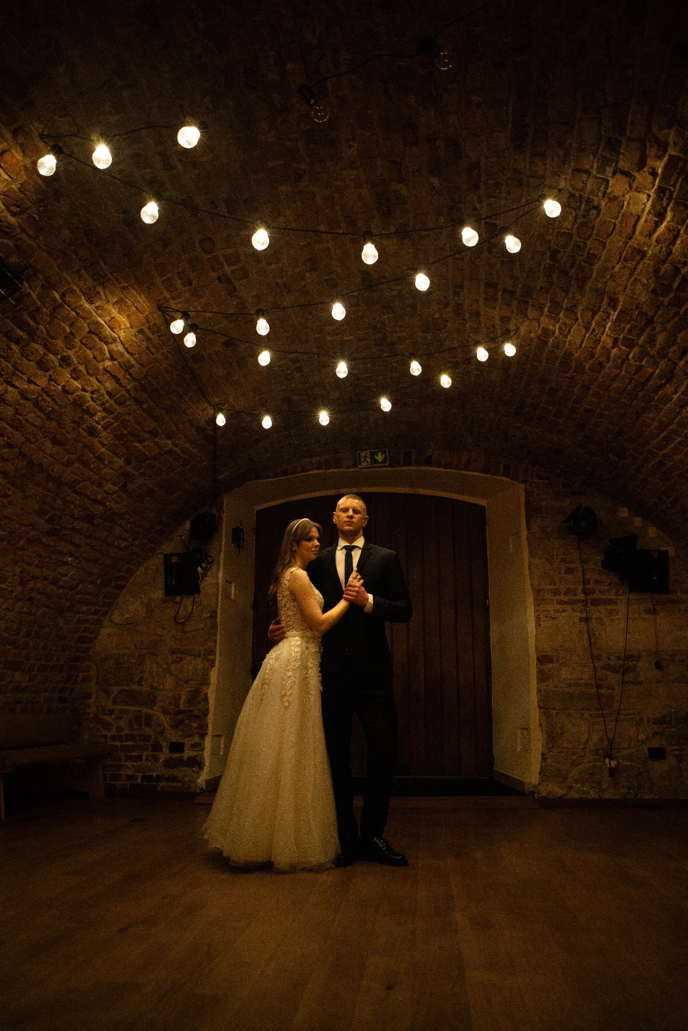 Para młoda w tańcu w ceglanym wnętrzu z dekoracyjnymi żarówkami. Panna młoda w sukni z koronką, pan młody w czarnym garniturze. Romantyczna atmosfera.