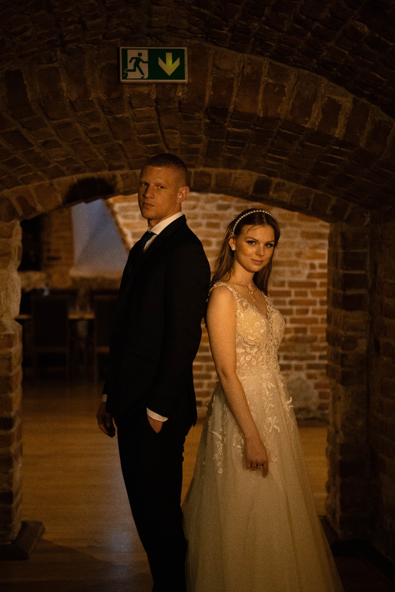 Para młoda w eleganckich strojach pozuje w ceglanym tunelu z zielonym znakiem ewakuacyjnym. Ona w sukni ślubnej, on w czarnym garniturze. Ciemne, nastrojowe oświetlenie.