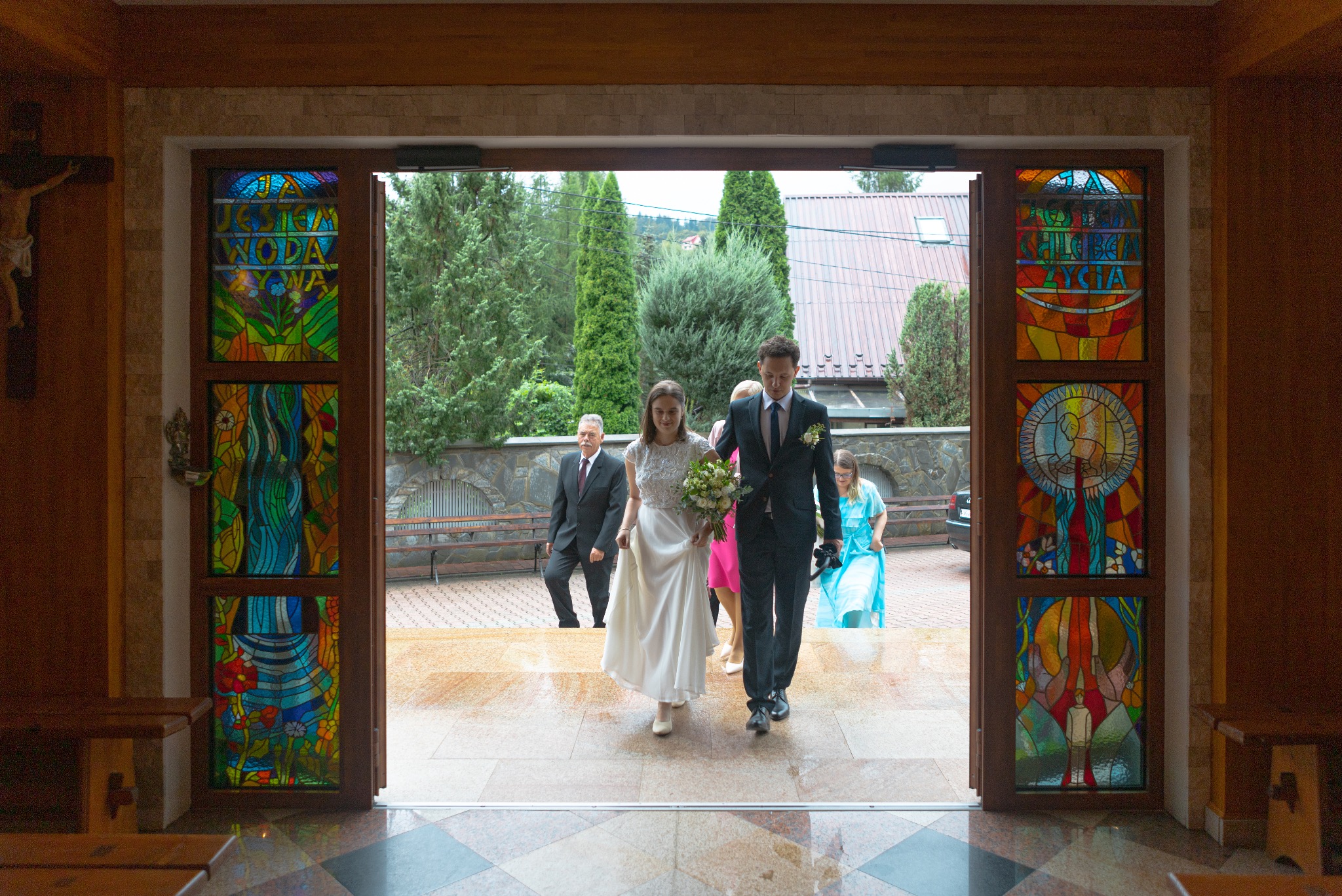 Para młoda wychodzi z kościoła przez zdobione witrażami drzwi. W tle goście weselni i zieleń. Ujęcie z wnętrza kościoła, z widocznym fragmentem ławki.