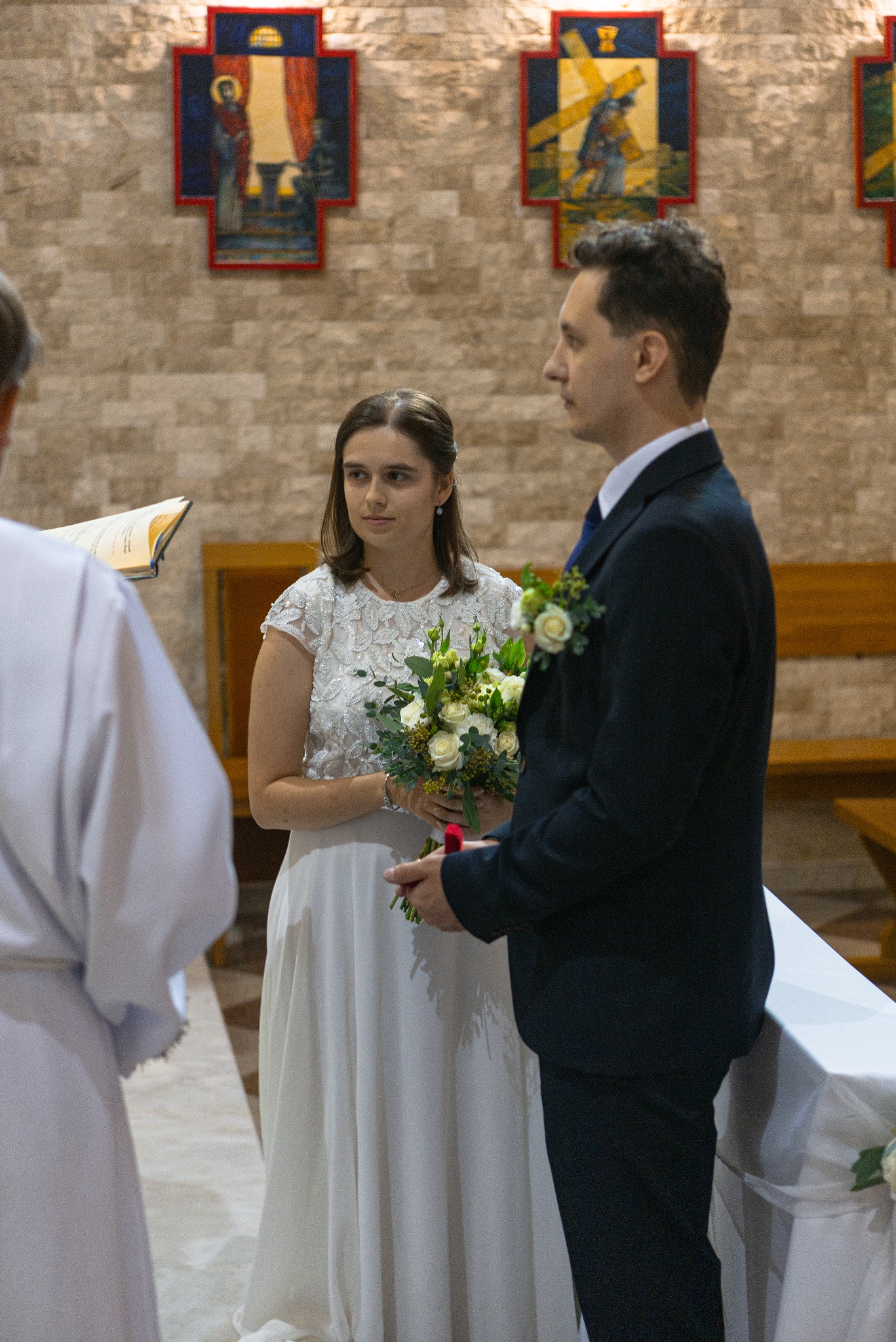 Para młoda podczas ceremonii ślubnej w kościele, panna młoda trzyma bukiet, w tle obrazy religijne na ceglanej ścianie.