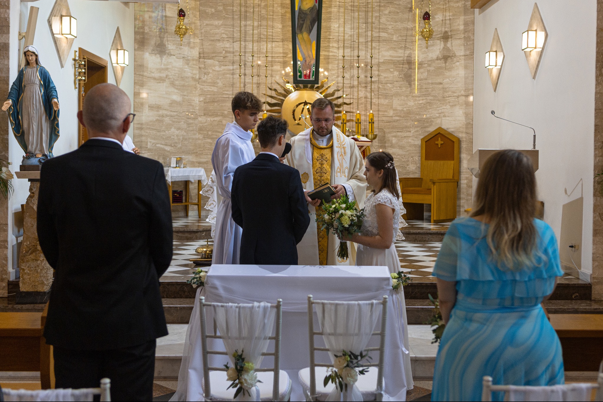 Uroczystość ślubna w kościele: para młoda przed księdzem, świadkowie i goście. Dekoracje kwiatowe na krzesłach i ołtarzu. Ciepłe oświetlenie podkreśla sakralny charakter.