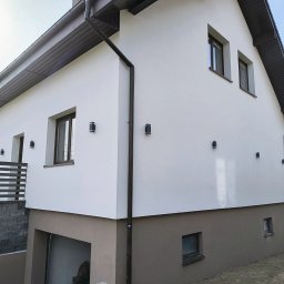 Dwukondygnacyjny dom z białą elewacją i brązowym cokołem, oświetlony zewnętrznymi lampami. Widoczny fragment dachu i rynny. Ujęcie z dołu, podkreślające geometrię budynku.