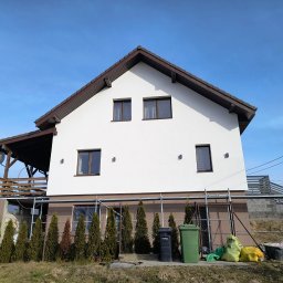 Dwukondygnacyjny dom z białą elewacją i brązowym dachem, otoczony rusztowaniami i zielonymi tujami. Widoczne worki z materiałami budowlanymi i kosze na śmieci.