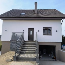 Nowoczesny dom jednorodzinny z białą elewacją, brązowym dachem i garażem. Widoczne schody wejściowe z metalową barierką i elementy wykończeniowe.