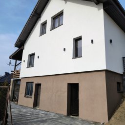 Dwukondygnacyjny dom z białą elewacją i brązowym cokołem, widoczny z poziomu chodnika z kostki brukowej. Uwagę zwracają nowoczesne lampy elewacyjne.