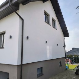 Julia bud - Dwukondygnacyjny dom z białą elewacją i brązowym cokołem, oświetlony lampami ściennymi. Widoczne okna, rynna i fragment dachu. Ujęcie z boku budynku.