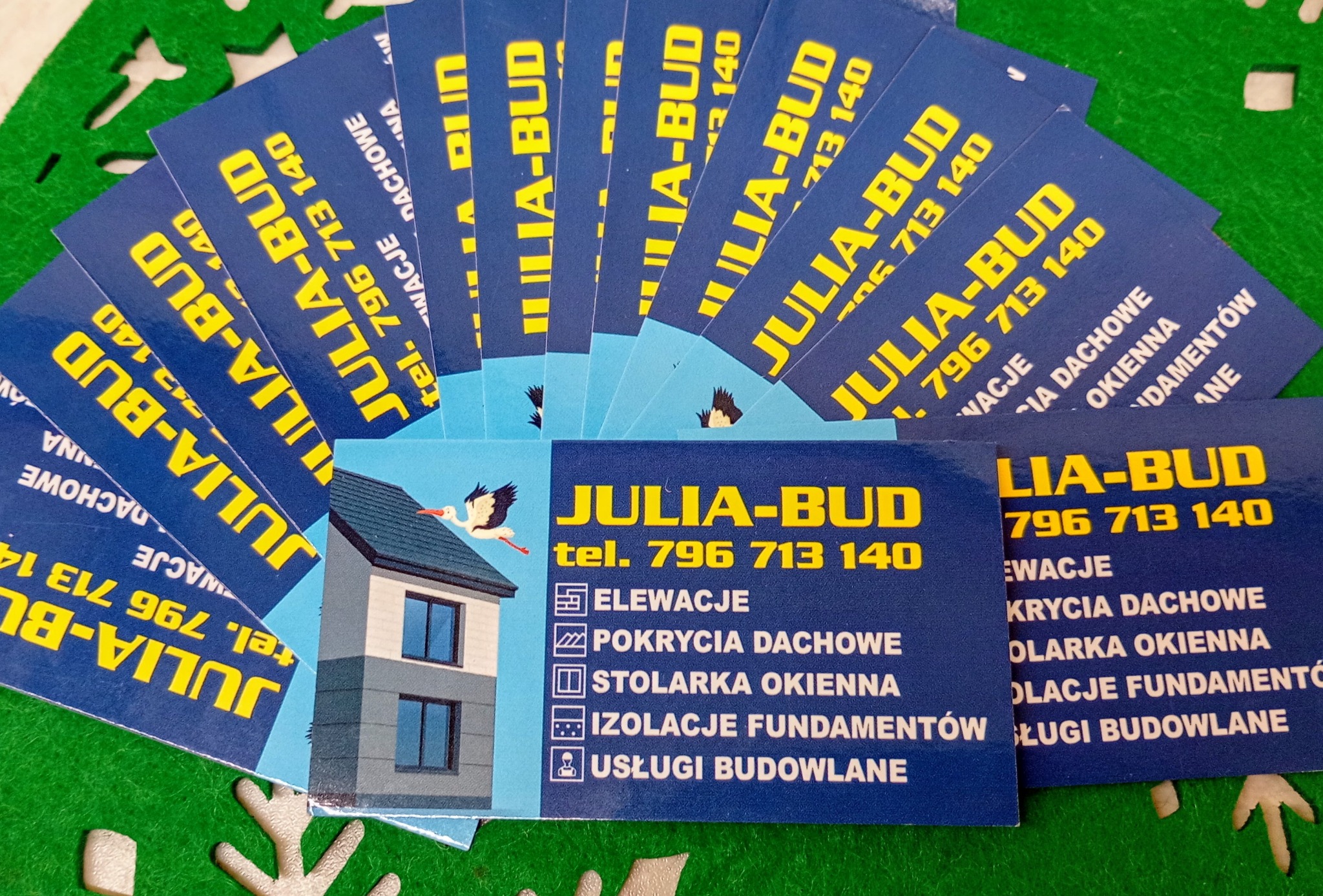 Wizytówki firmy Julia-Bud z ofertą: elewacje, pokrycia dachowe, stolarka okienna, izolacje fundamentów i usługi budowlane. Na wizytówce ilustracja domu i bocian.
