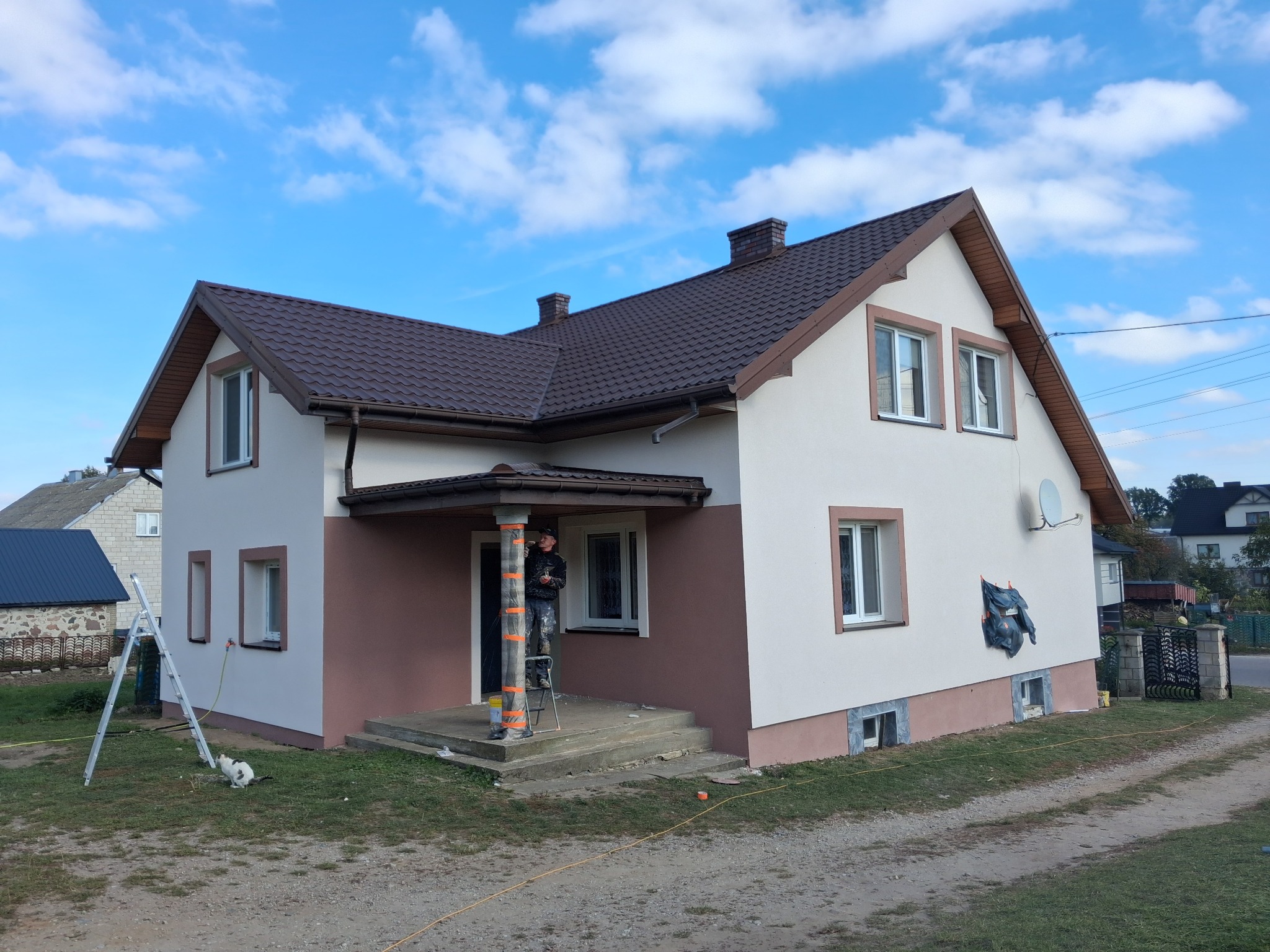 Dwukolorowa elewacja domu jednorodzinnego w trakcie malowania. Pracownik na drabinie zabezpiecza kolumnę wejściową. Widoczne rusztowanie i akcesoria malarskie.