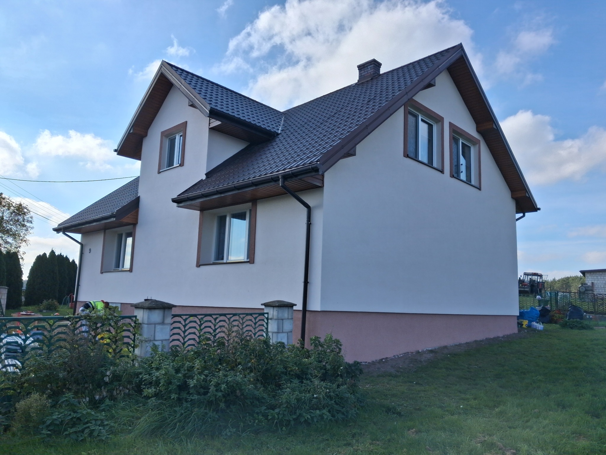 Dwukondygnacyjny dom z ciemnym dachem i jasną elewacją, widoczny z boku, z zielonym trawnikiem i częściowo widocznym ogrodzeniem. Błękitne niebo z chmurami w tle.