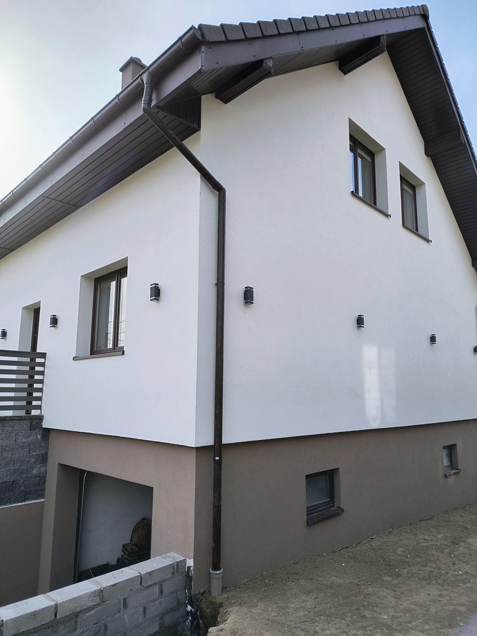 Dwukondygnacyjny dom z białą elewacją i brązowym cokołem, oświetlony zewnętrznymi lampami. Widoczny fragment dachu i rynny. Ujęcie z dołu, podkreślające geometrię budynku.