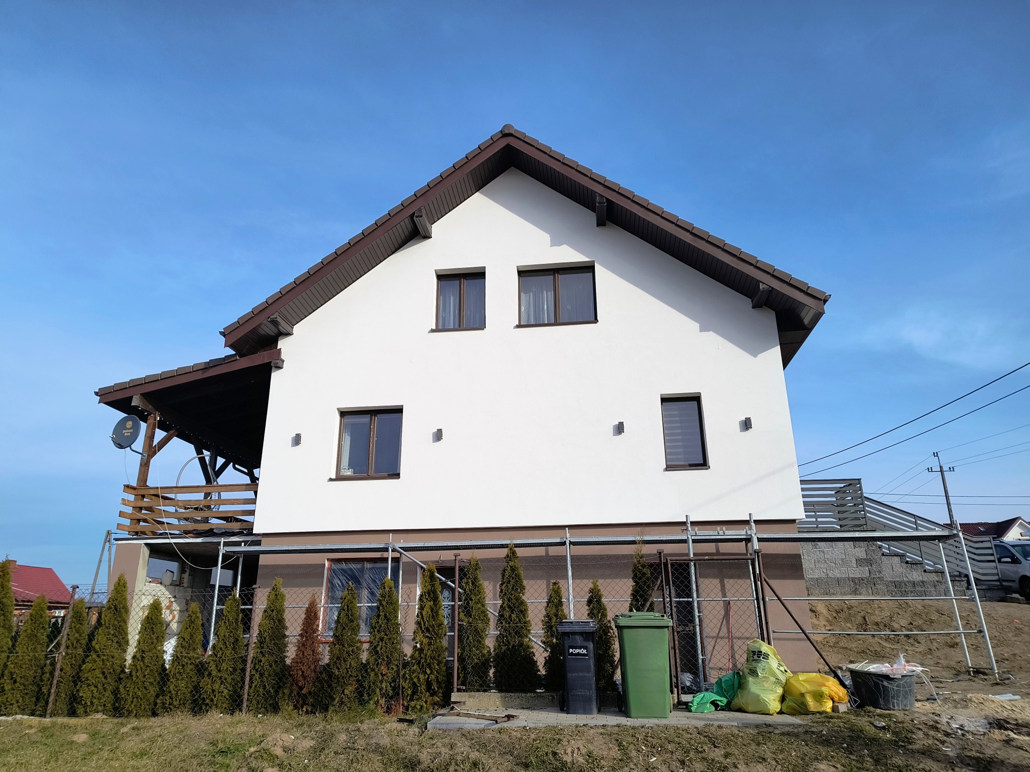 Dwukondygnacyjny dom z białą elewacją i brązowym dachem, otoczony rusztowaniami i zielonymi tujami. Widoczne worki z materiałami budowlanymi i kosze na śmieci.