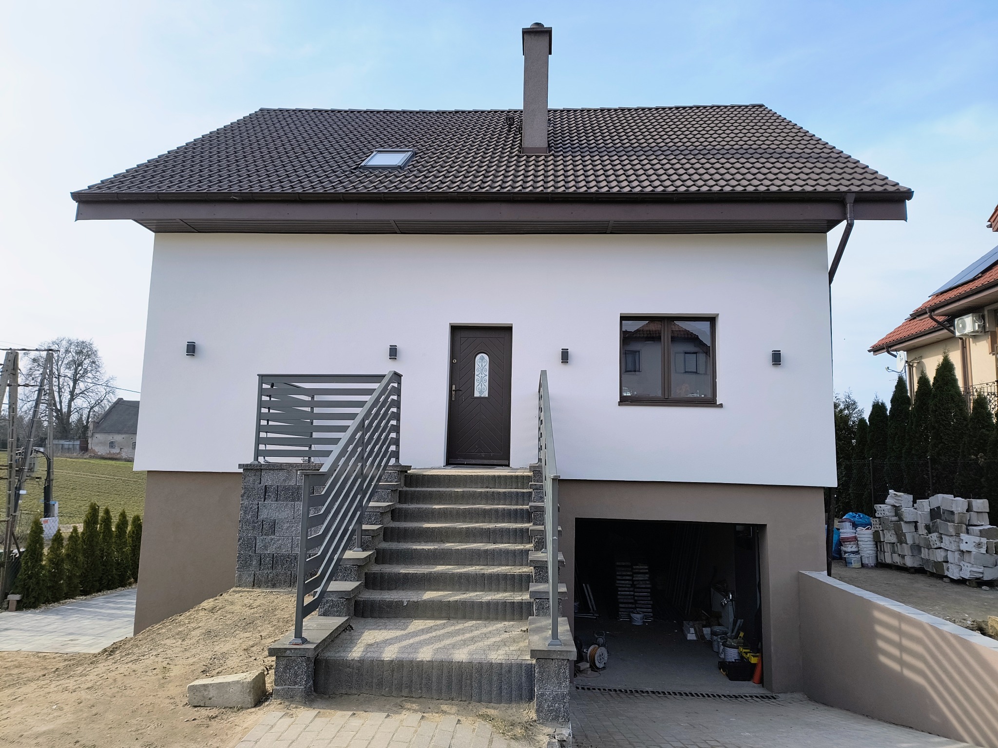 Nowoczesny dom jednorodzinny z białą elewacją, brązowym dachem i garażem. Widoczne schody wejściowe z metalową barierką i elementy wykończeniowe.