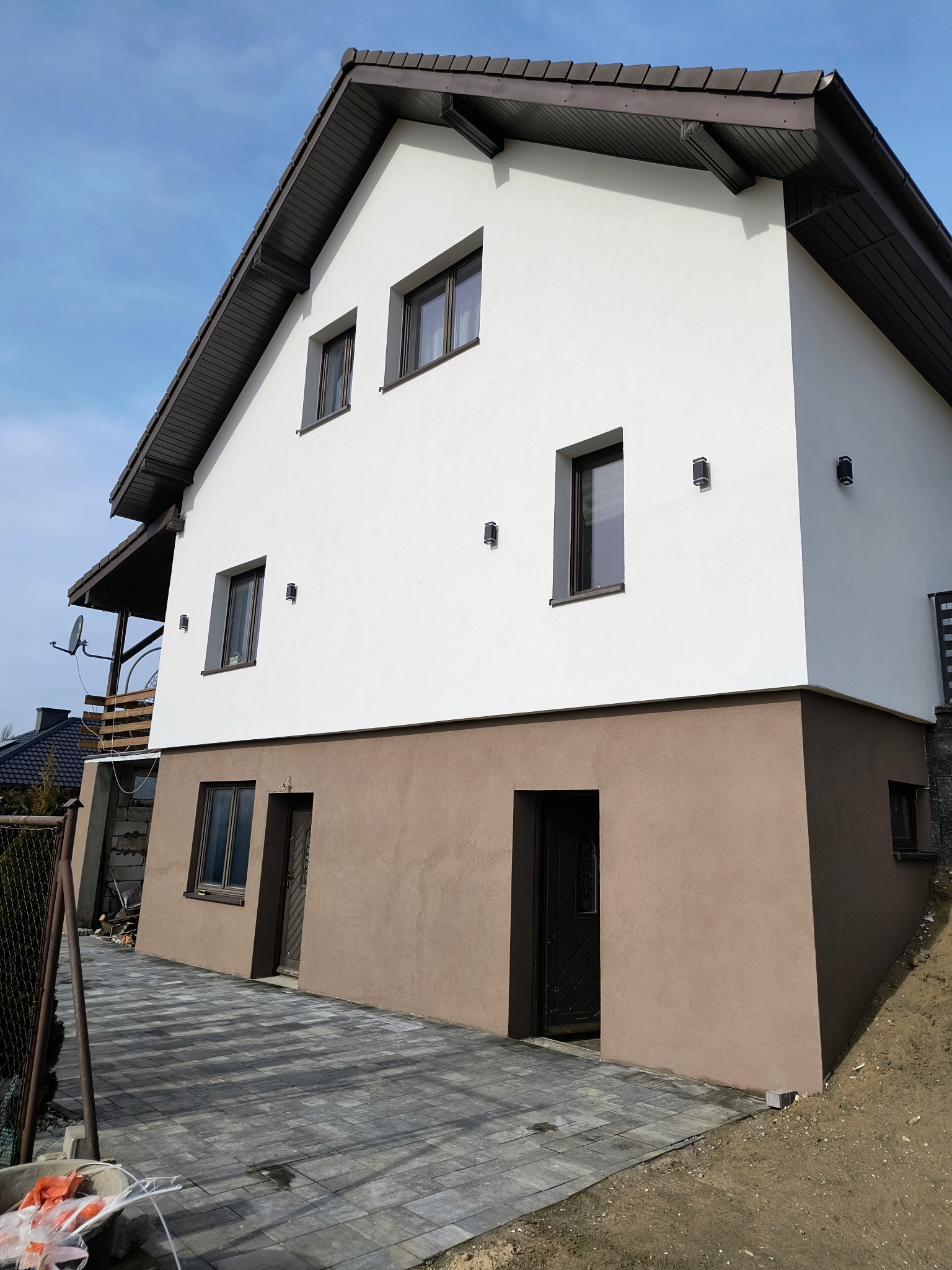 Dwukondygnacyjny dom z białą elewacją i brązowym cokołem, widoczny z poziomu chodnika z kostki brukowej. Uwagę zwracają nowoczesne lampy elewacyjne.