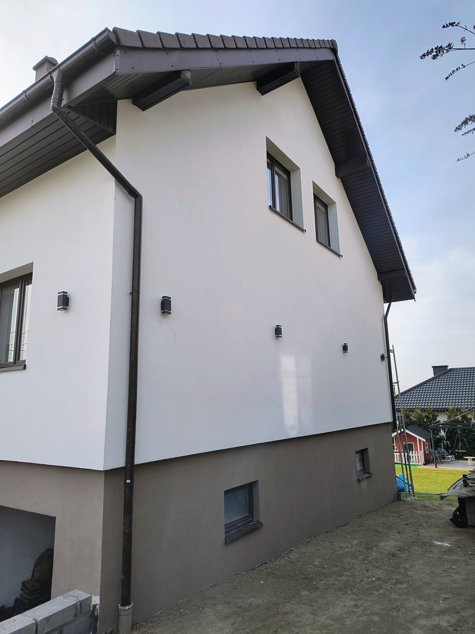 Dwukondygnacyjny dom z białą elewacją i brązowym cokołem, oświetlony lampami ściennymi. Widoczne okna, rynna i fragment dachu. Ujęcie z boku budynku.