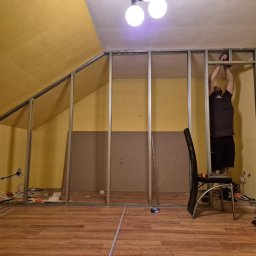 KOZ-BUD Mateusz Kozłowski - Montaż metalowej konstrukcji ścianki działowej w pokoju na poddaszu, widoczny pracownik, narzędzia i częściowo zamontowana płyta. Żółte ściany i drewnopodobna podłoga.