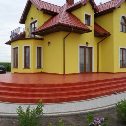 Koza Buduje - Żółty dom z czerwoną dachówką i schodami, widok z dołu. Balustrada balkonu, lampy zewnętrzne i ozdobne okna dodają uroku. W tle zielona roślinność i samochód.