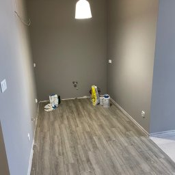 KOZ-BUD Mateusz Kozłowski - Szare wnętrze w trakcie malowania: widoczne puszki z farbą, worki z zaprawą i drewnopodobna podłoga. Świeżo pomalowane ściany i sufit podwieszone.