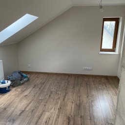KOZ-BUD Mateusz Kozłowski - Poddasze po malowaniu ścian na neutralny odcień, z drewnianą podłogą, oknem dachowym i pionowym. Widoczne akcesoria malarskie i grzejnik.
