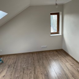 KOZ-BUD Mateusz Kozłowski - Poddasze po malowaniu w Gostyninie. Widoczne okno dachowe, drewniana podłoga i świeżo pomalowane ściany na biało. Częściowo widoczne materiały malarskie.