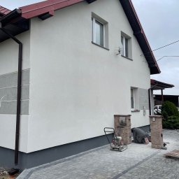 KOZ-BUD Mateusz Kozłowski - Dwukondygnacyjny dom z jasną elewacją i czerwoną dachówką. Ułożona kostka brukowa, widoczny pracownik i zagęszczarka. Detal: dekoracyjne wykończenie elewacji.