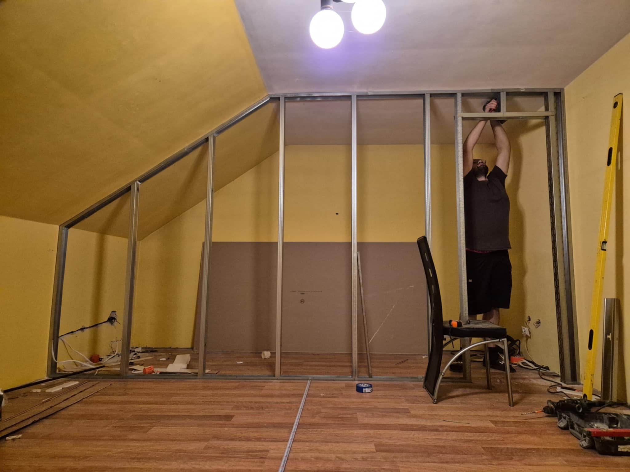 Montaż metalowej konstrukcji ścianki działowej w pokoju na poddaszu, widoczny pracownik, narzędzia i częściowo zamontowana płyta. Żółte ściany i drewnopodobna podłoga.