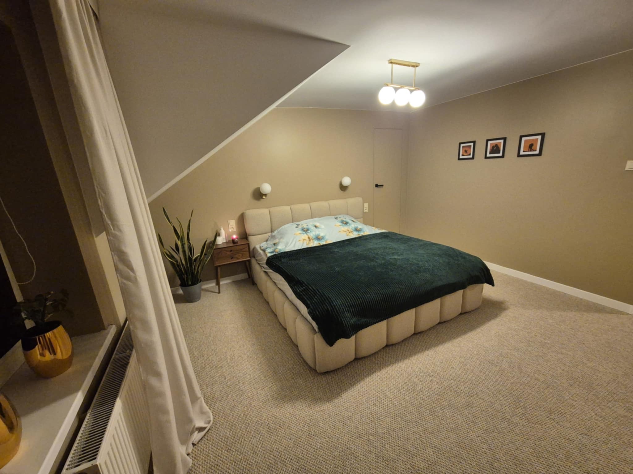 Beżowa sypialnia na poddaszu z łóżkiem typu boxspring, zielonym pledem i trzema obrazkami na ścianie. Widoczny fragment okna z zasłoną i grzejnikiem.