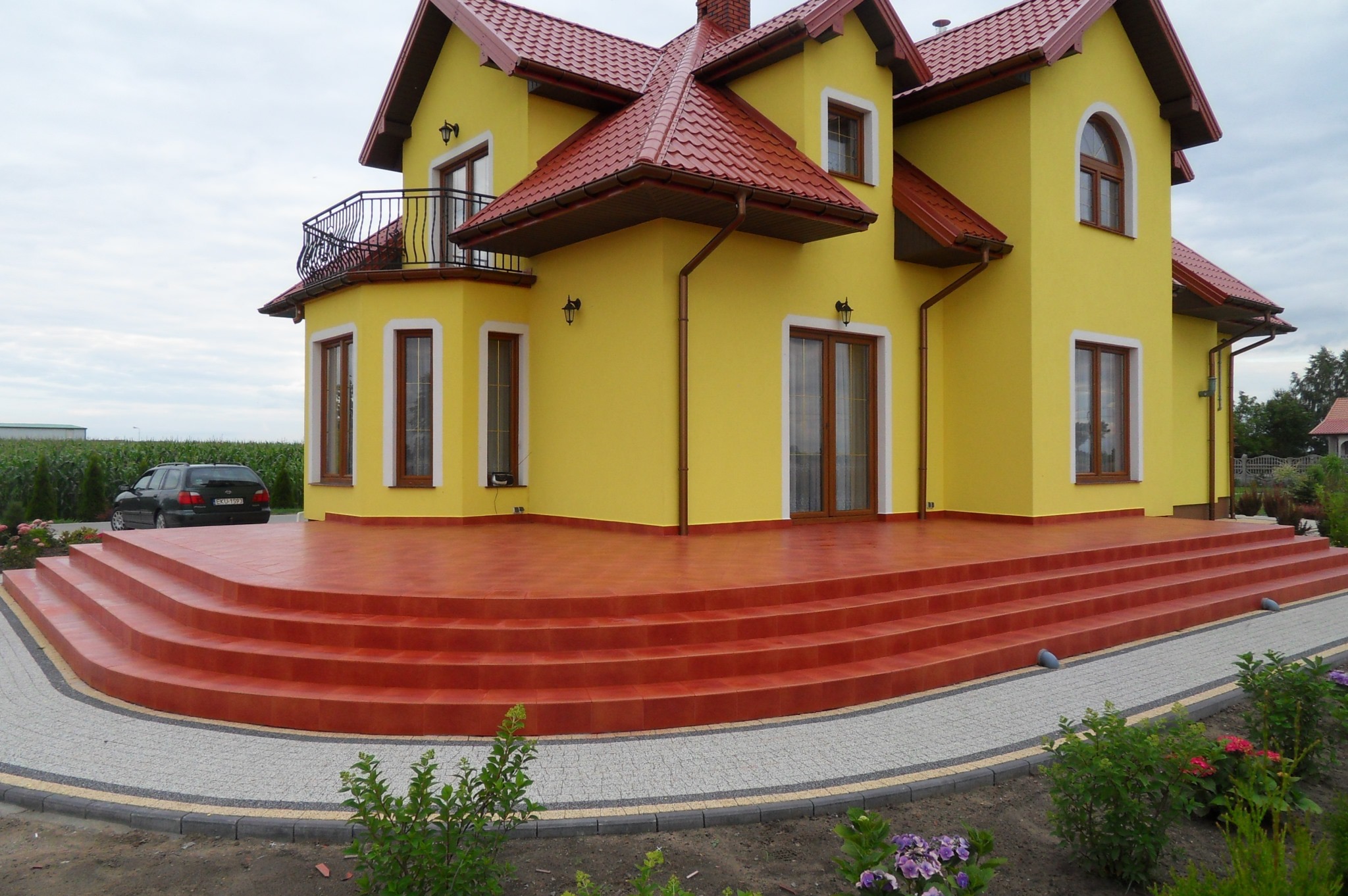 Żółty dom z czerwoną dachówką i schodami, widok z dołu. Balustrada balkonu, lampy zewnętrzne i ozdobne okna dodają uroku. W tle zielona roślinność i samochód.